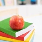 Three school books stacked with an apple on top in a classroom