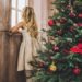 a blonde girl looking out the window by a Christmas tree